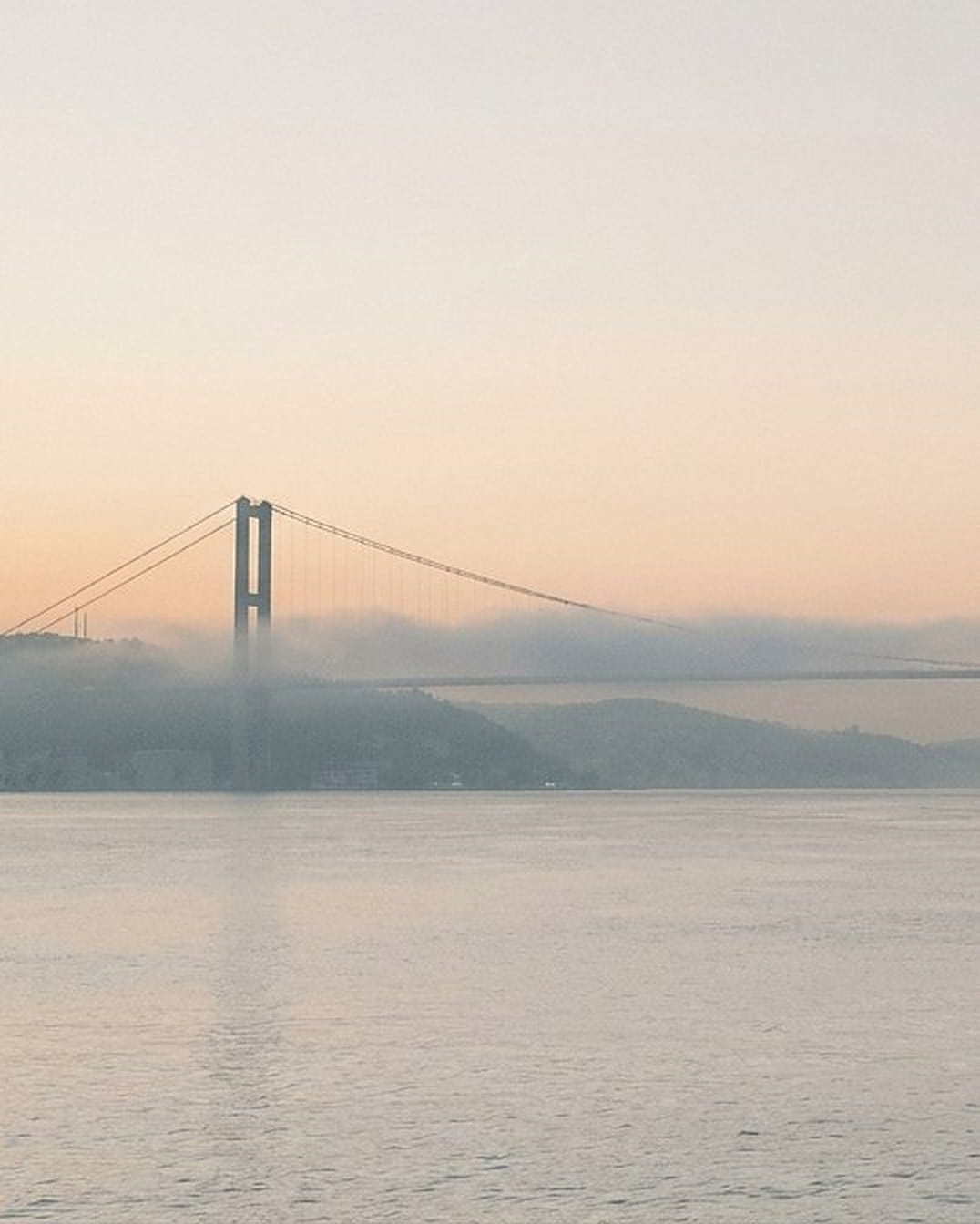 Hängebrücke im Morgendunst über breitem, ruhigem Wasser, Hügel an beiden Ufern